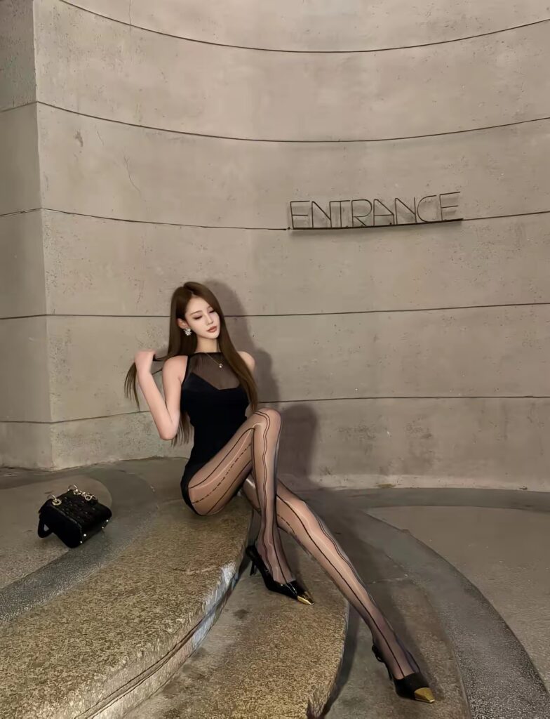 弧形石墙“ENTRANCE”标识处,长发女生穿黑色包臀小礼服与波纹条纹透肤连裤袜,坐在石阶上伸腿摆拍,左侧放着黑色手提包,尖头高跟带金属装饰。