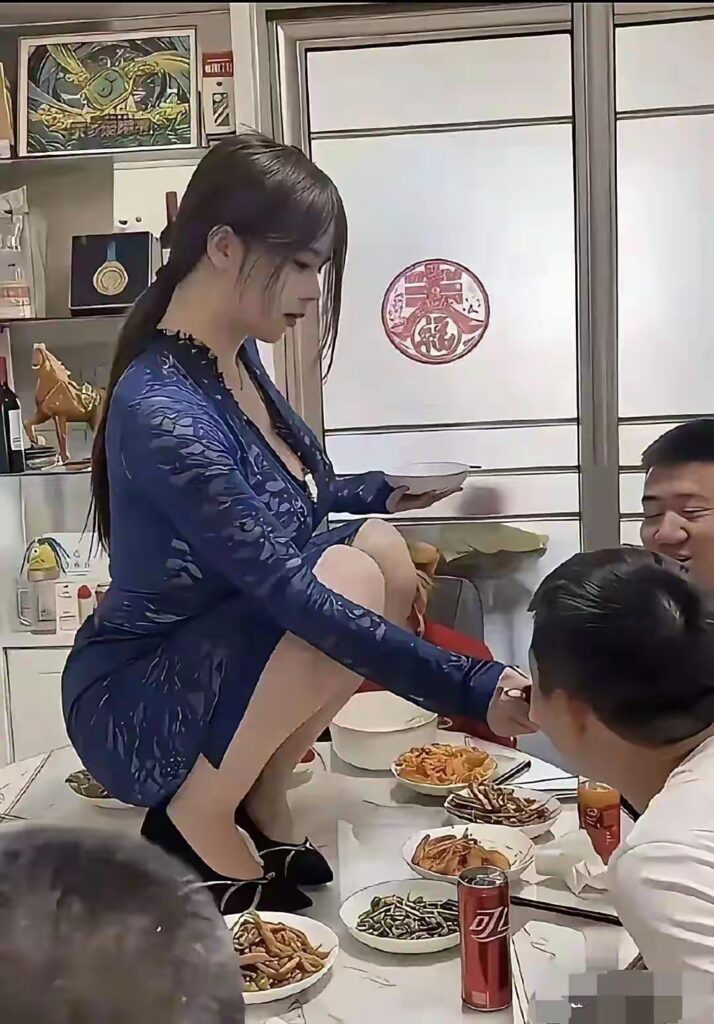 同一聚餐环境的近景，蓝色蕾丝裙女子蹲在餐桌上，手持小碗并伸手与右侧男士互动，桌面菜肴丰富，饮料与纸巾可见。