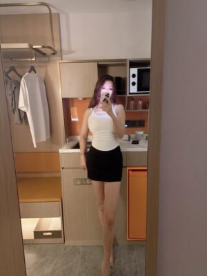 现代酒店客房内，一位女子穿白色吊带上衣与黑色短裙，对镜全身自拍；背景有小型吧台、微波炉、挂衣区与橙色抽屉，暖色灯光。