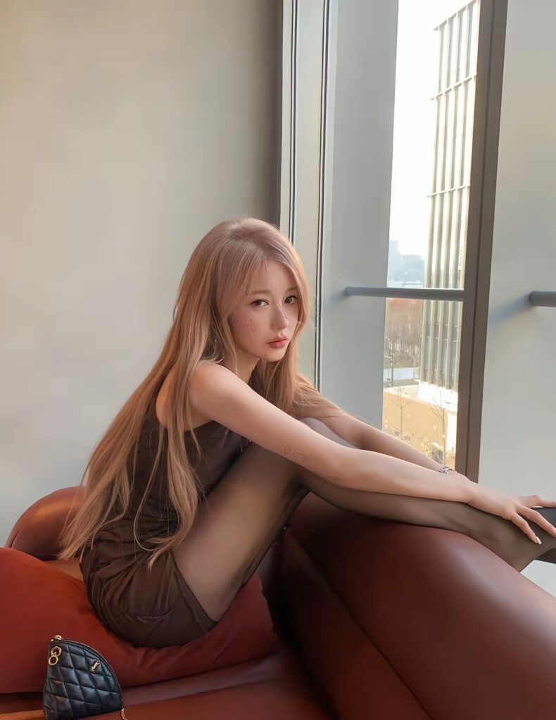 女生在落地窗前侧坐，长发披肩，穿咖色无袖连衣裙与黑色连裤袜，抱膝望向镜头。
