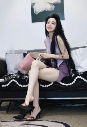 女生坐在沙发边笑看镜头，双腿交叠，一只鞋半脱挂在脚尖；紫色上衣与包臀短裙同色，周围有购物袋。