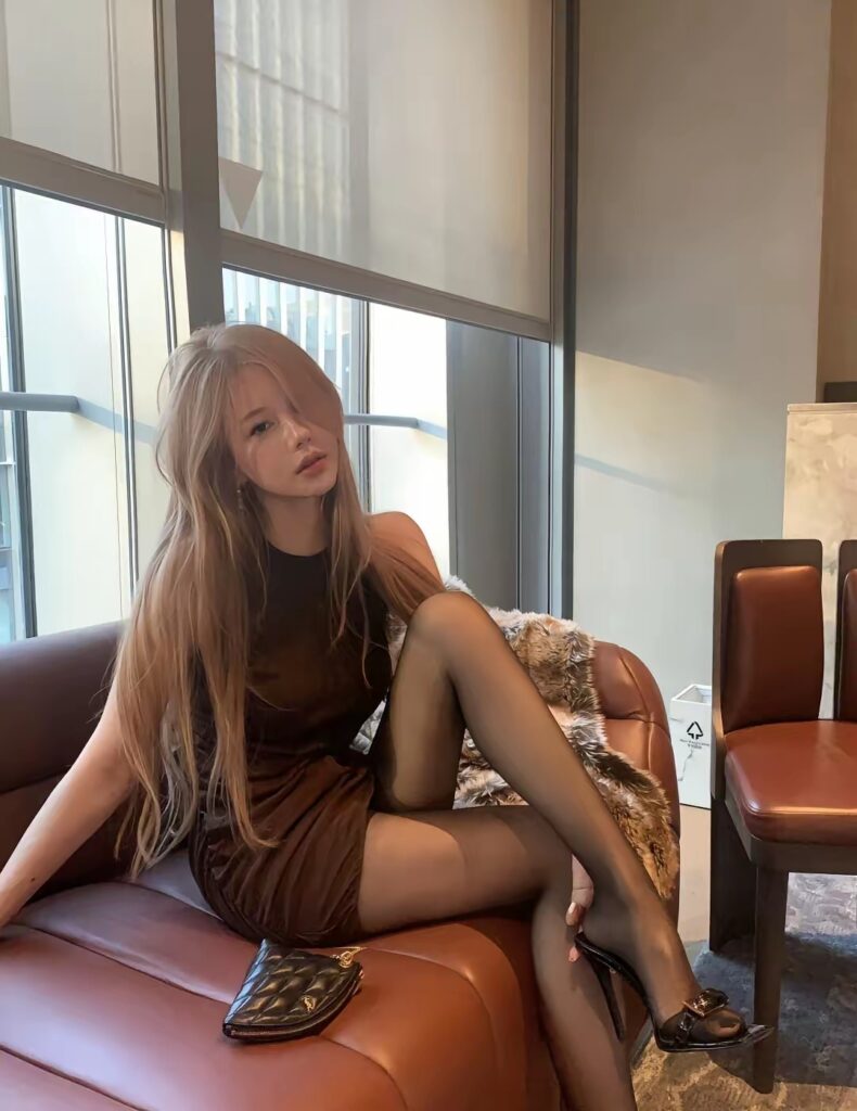 女生坐在沙发扶手处，咖色连衣裙与黑色连裤袜搭配，脚穿黑色高跟鞋，身后是毛绒披毯，小菱格包置于座面。