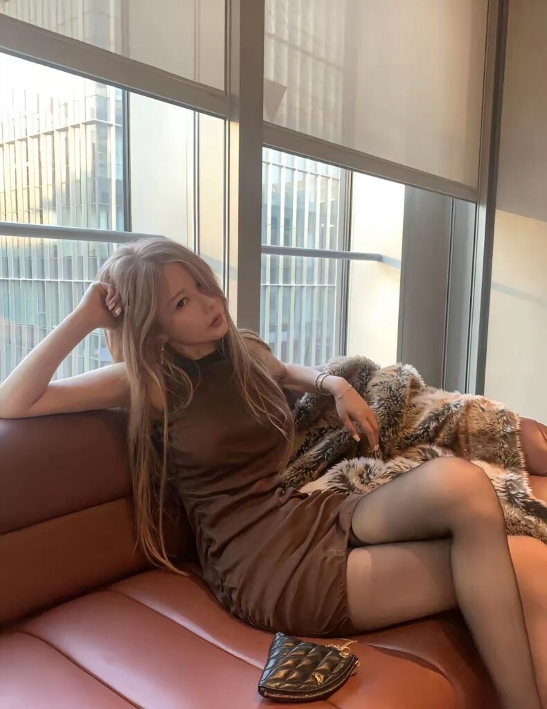 女生半躺在沙发上，咖色连衣裙配黑色连裤袜，手扶头靠，旁边铺着毛绒披毯与小号菱格包。