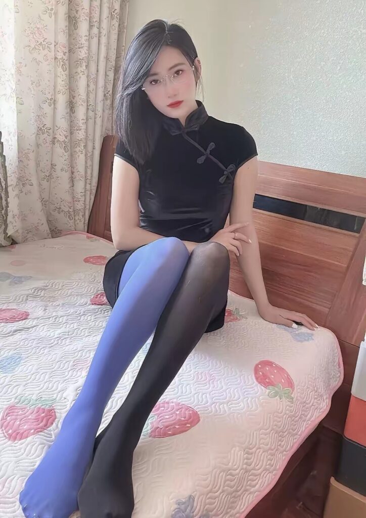 坐在床边的女子穿黑色旗袍，左右腿蓝黑拼色连裤袜，微挑下巴直视镜头