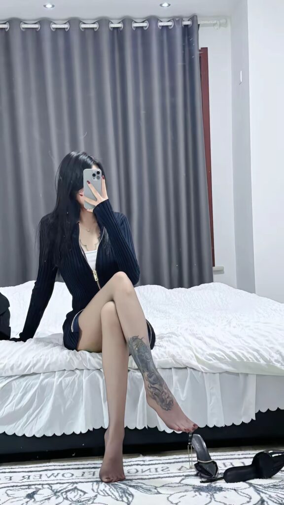 女生端坐床沿回看镜中，左脚踢起一只高跟鞋，连裤袜与纹身腿自然垂落，床下散落另一只鞋。