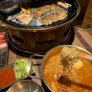 圆形铁板烤炉上正烤着腌肉、五花肉和小香肠，旁边铝杯里是汤汁；前景是一只黄铜小锅的辣味泡面，筷子正夹起面条，桌上配有蘸酱与青椒圈。