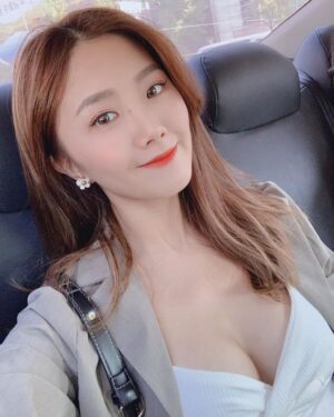 车内光线柔和，一位女生微笑自拍，佩戴小白花耳饰，妆容清爽，身穿浅灰外套与白色V领上衣，肩背黑色细带包。