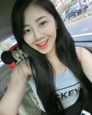 车内自然光下的女生微笑自拍，长发、灰色背心，靠枕为卡通图案；窗外车流与道路虚化，画面轻松明亮。