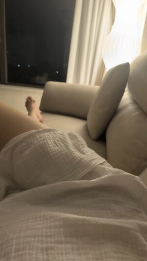 夜晚的酒店客房里，暖黄色立灯照亮米色沙发，一人穿浅色浴袍侧躺放松，窗外可见零星灯点，氛围安静克制。