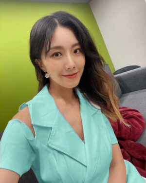 室内绿墙背景下，女生穿薄荷绿露肩连衣裙，侧披长发，对镜头微笑，沙发上有红色毯子。