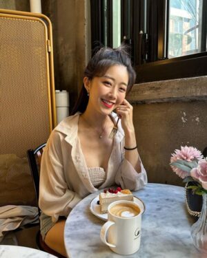 阳光下的咖啡馆里，女生穿浅色衬衫外搭与短上衣，面前摆着拿铁与奶油蛋糕，对镜微笑。