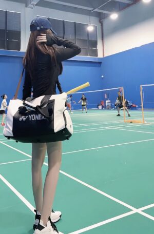 羽毛球馆内，一位女生背着YONEX球包、戴帽子站在场边观看。