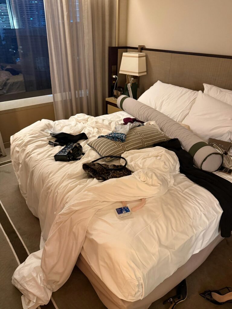 夜晚的酒店房间，床上堆满衣物与包包，床边与地上散落高跟鞋。