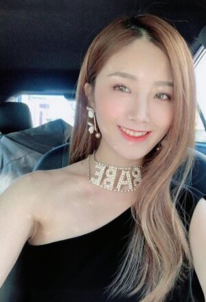 车内自然光下的女生微笑自拍，穿黑色单肩上衣，佩戴珍珠字母项链与耳饰，背景为后座与靠枕。