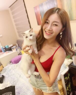 室内暖光下，女生穿红色吊带与牛仔短裤怀抱白猫对镜微笑，背景是卧室床与日常杂物，氛围轻松。