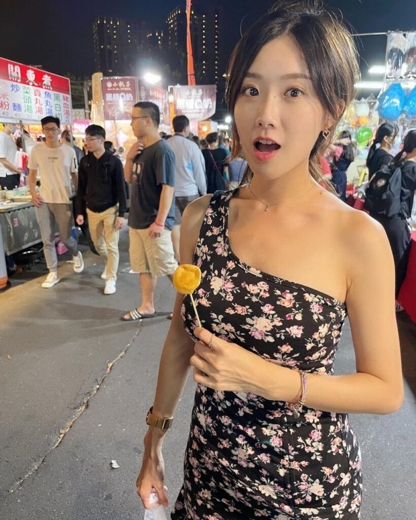 夜市灯光下，穿黑底碎花单肩连衣裙的女生微笑看镜头，手举一串金黄小吃，周围人潮热闹。